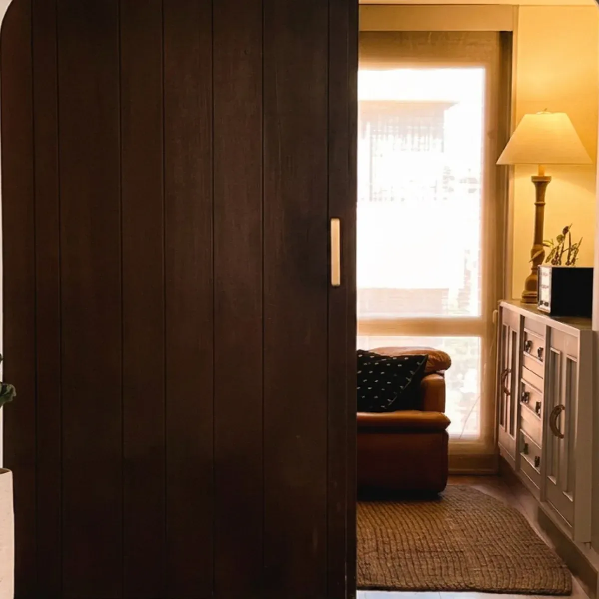 Puerta corrediza de madera oscura entreabierta hacia una sala cálida con sofá de cuero y una lámpara encendida