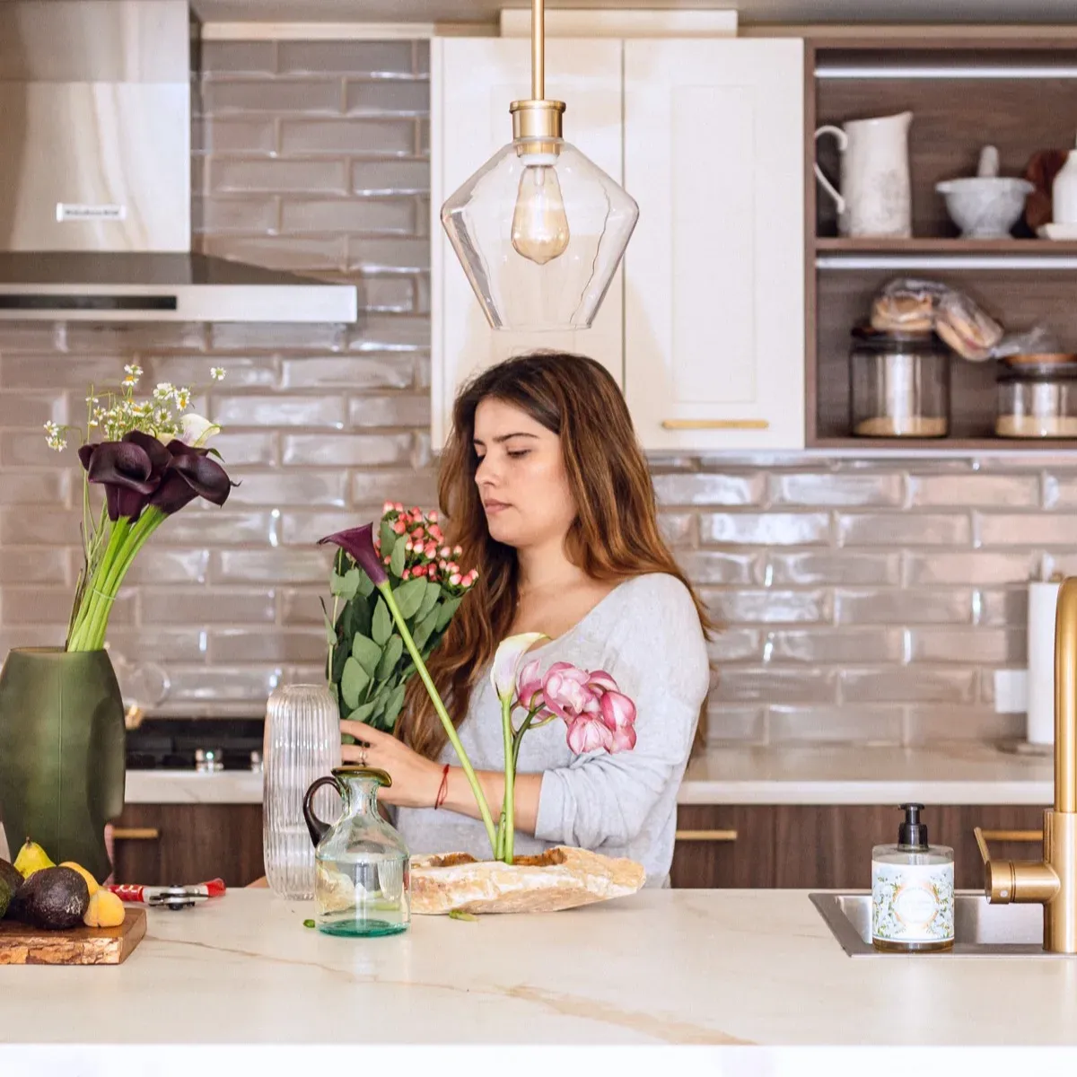 Carolina arreglando un ramo de flores sobre la isla de una cocina con azulejos y lámpara de vidrio