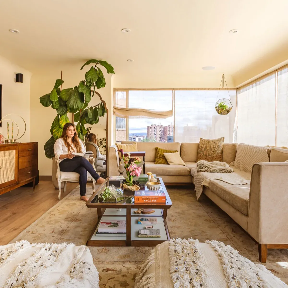 Mujer leyendo en un sillón junto a una planta grande, en una sala con sofá beige y luz natural